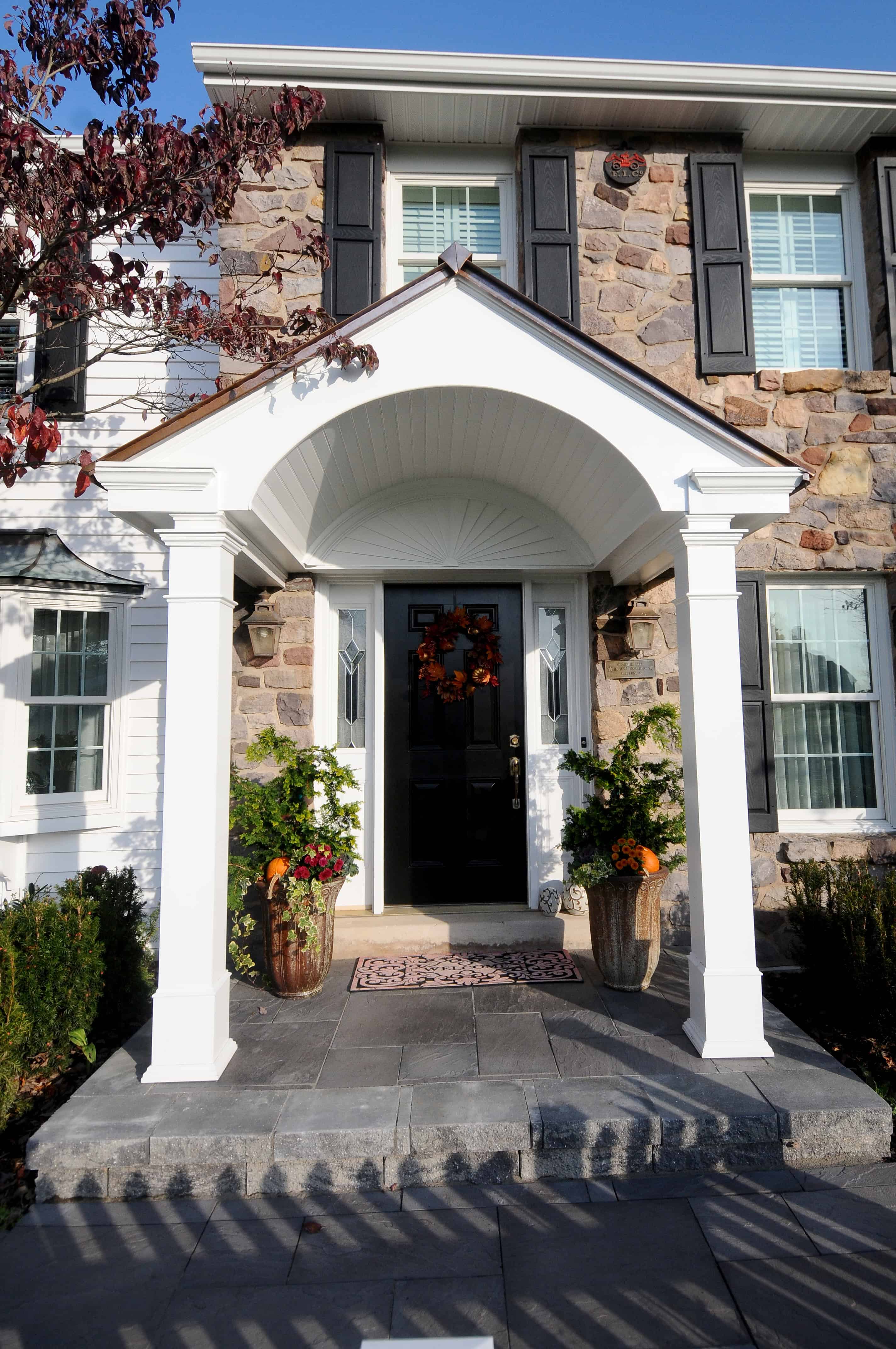 Front entrance of a stone house with white pillars, a covered porch, potted plants, and a dark front door decorated with a fall wreath. Steps lead up to the porch, and sunlight casts shadows on the walkway.