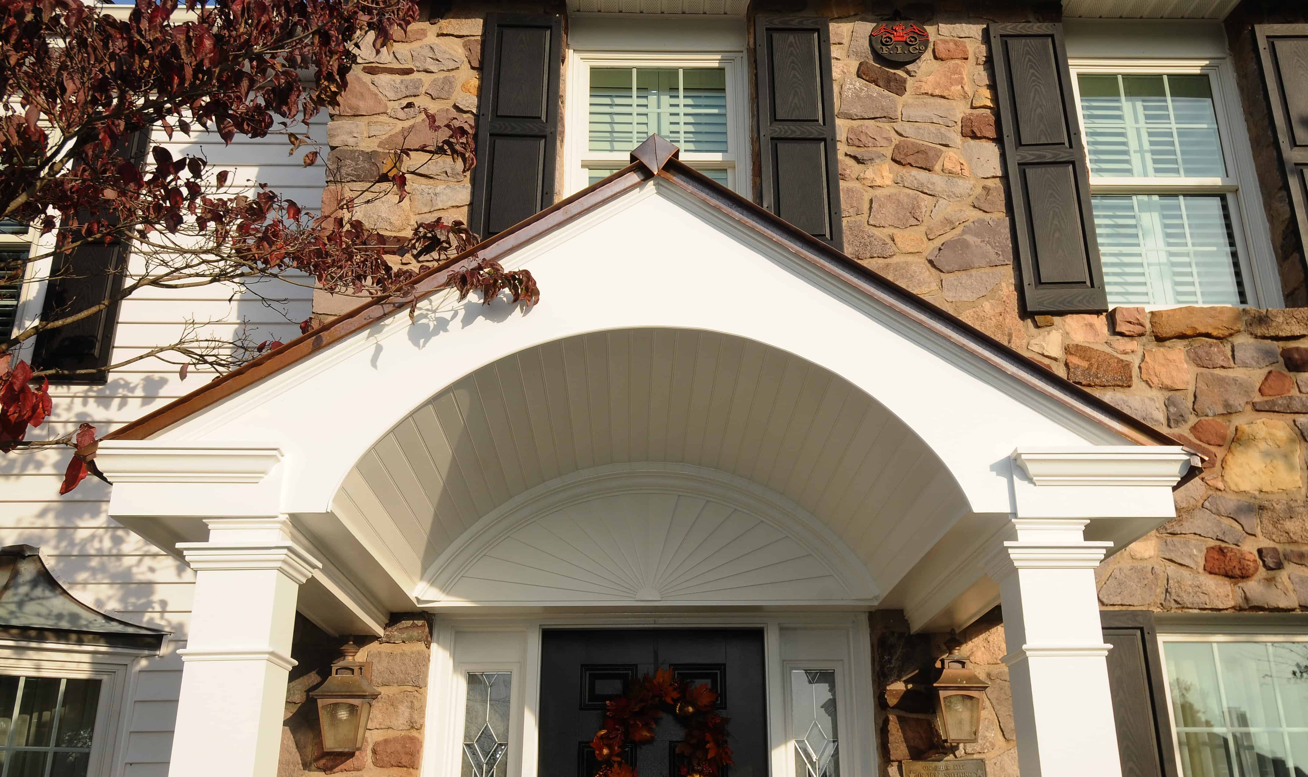 A close-up view of a house exterior with stone walls, black window shutters, and a white arched porch above the front door decorated with a fall wreath.