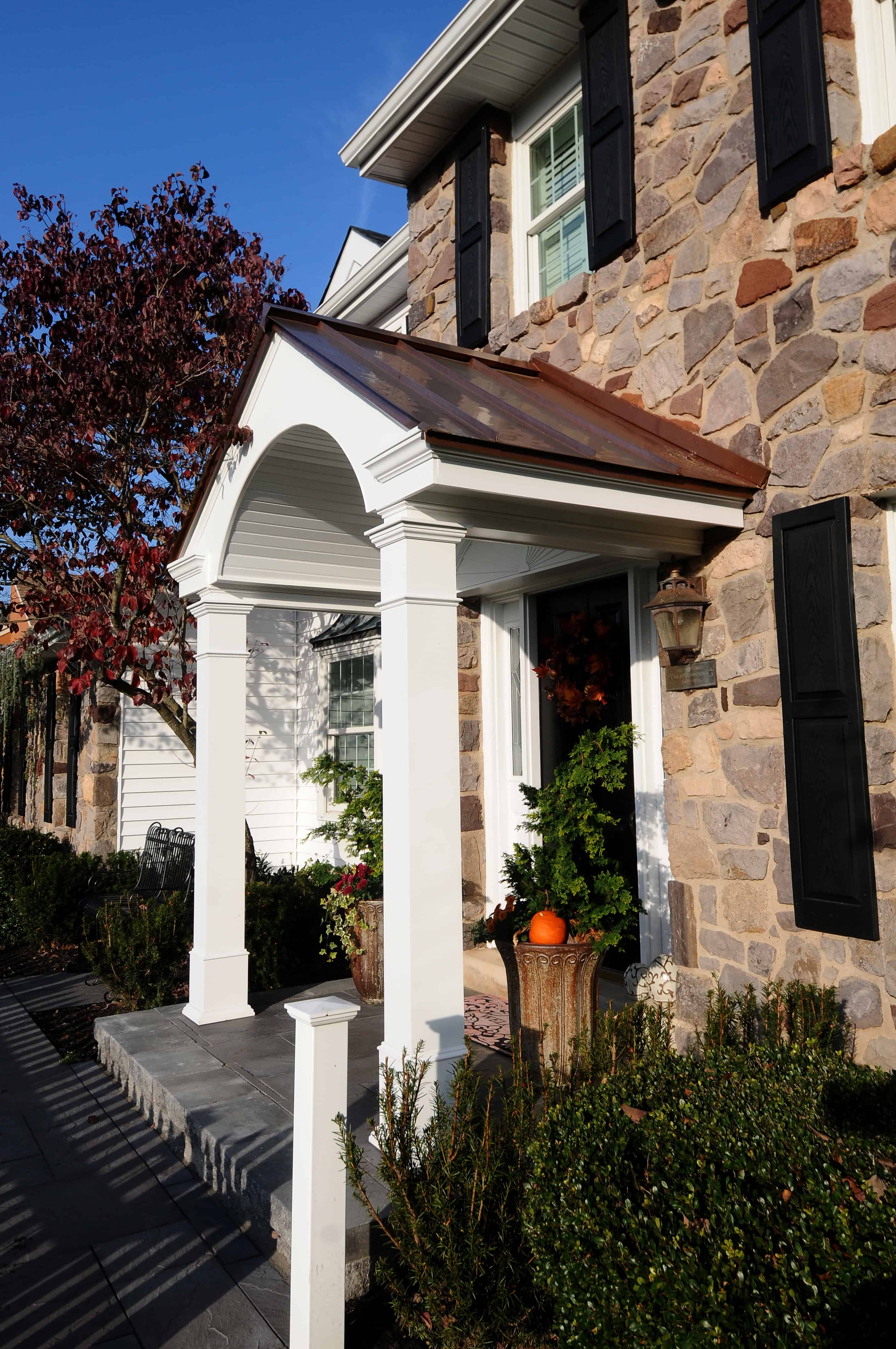 A stone-faced house with black shutters and a white columned porch. A potted plant and a pumpkin sit near the entrance, and a tree with red leaves is visible in the background under a clear blue sky.