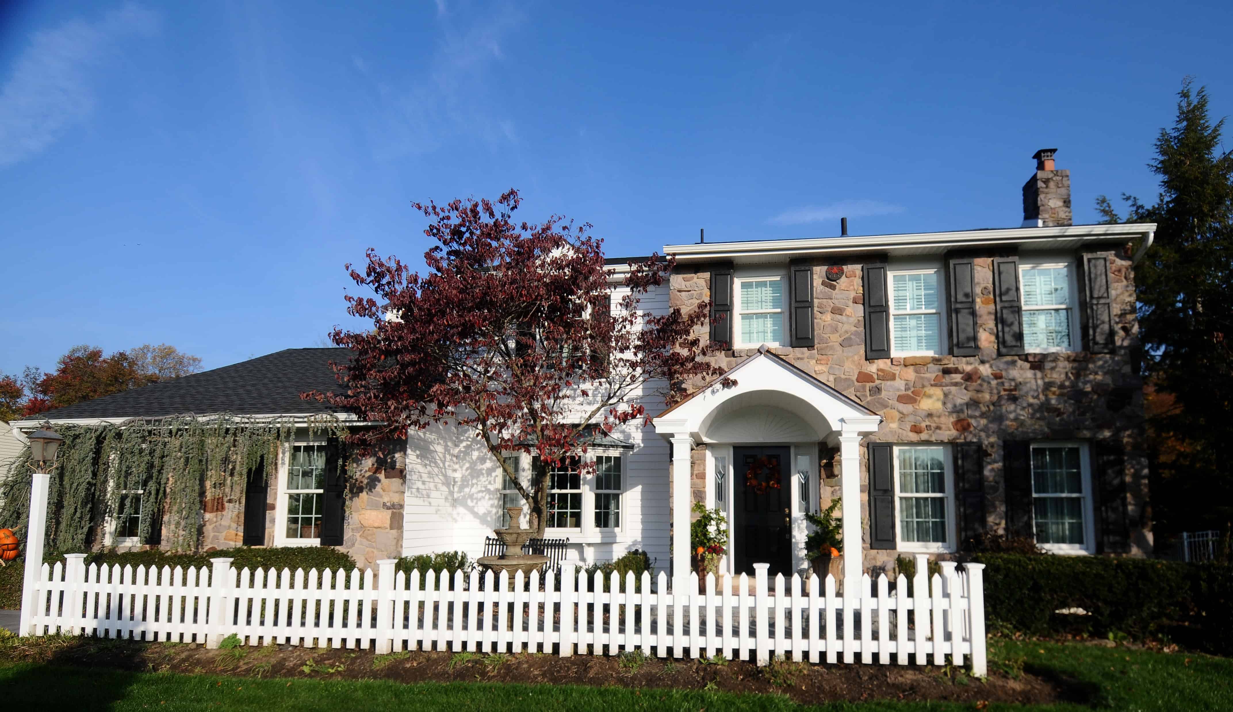 A two-story house with a stone facade, blue shutters, a white picket fence, and a front porch. There are trees and shrubs in the yard under a clear blue sky.