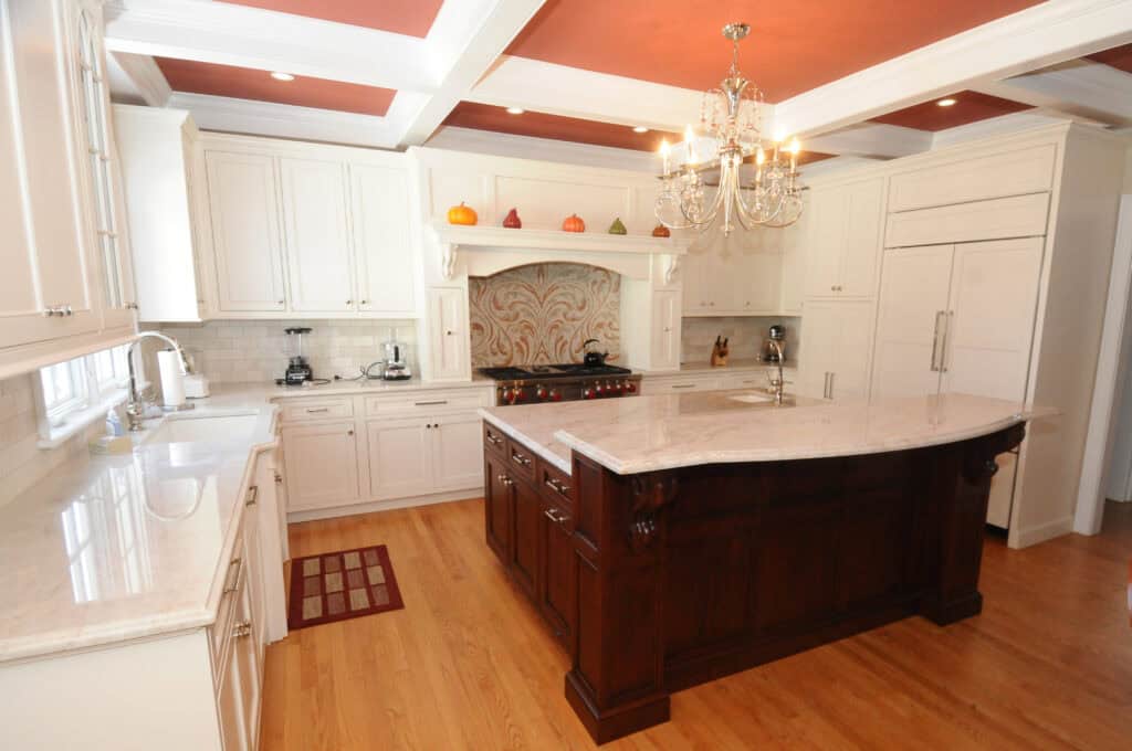 Spacious kitchen with white cabinets, a large dark wood island, chandelier, hardwood floors, autumn decorations above the stove, and built-in appliances. The ceiling is painted a warm reddish color with white beams.