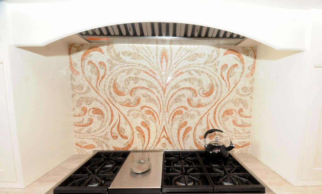 A kitchen stovetop with four gas burners, a central griddle, and a decorative tile backsplash with a swirling leaf pattern. A chef-shaped ceramic container sits on the right side of the stovetop.