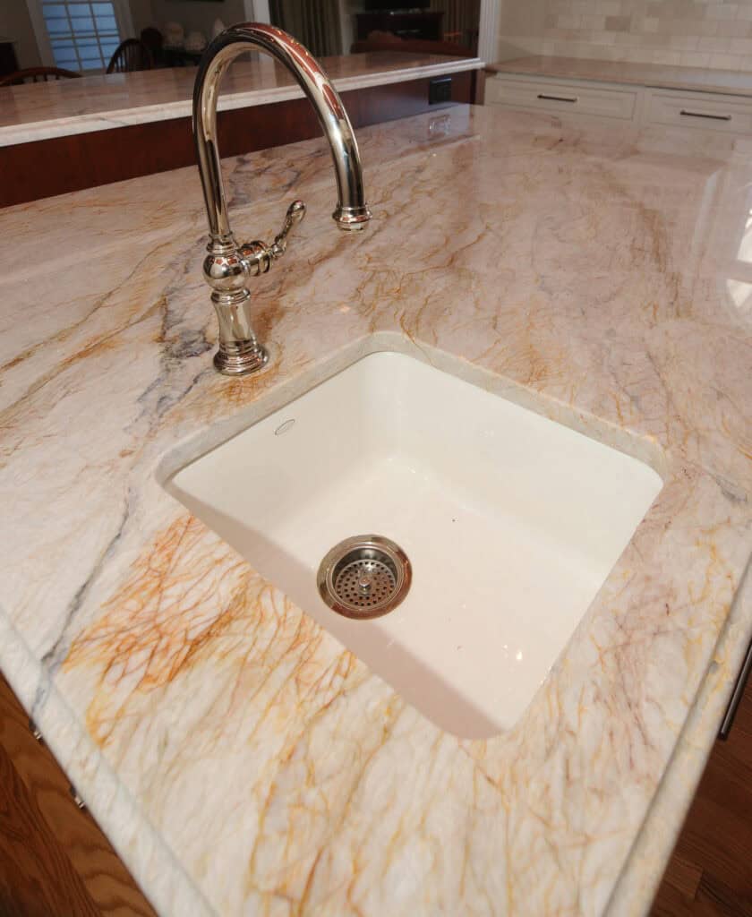 A close-up of a square white sink with a silver faucet set in a polished marble countertop with brown and gold veining, in a modern kitchen.