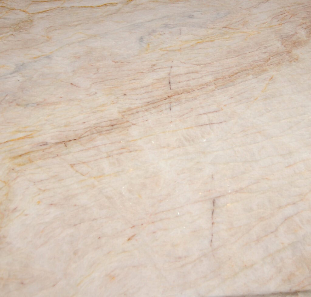 Light marble surface with subtle beige, cream, and tan tones, featuring natural veining and faint streaks of yellow and brown throughout. The texture appears smooth with minor variations in color.