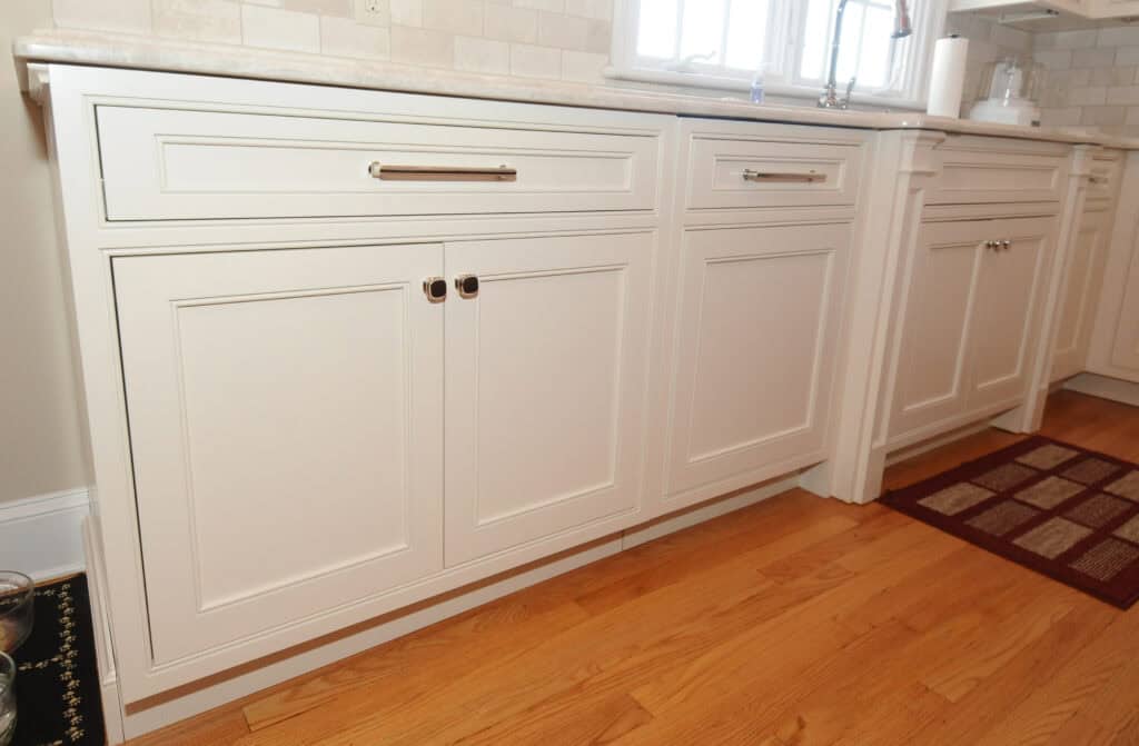 White kitchen cabinets with silver handles and a beige countertop, set against a light tiled backsplash. The cabinets are above a medium-toned wood floor, with a red and beige rug nearby. A sink is visible in the background.