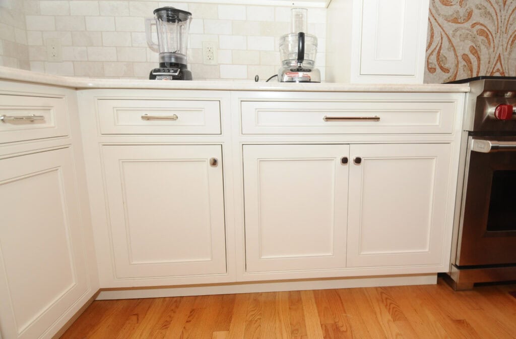 White kitchen cabinets with silver handles and dark round knobs are shown below a light-colored countertop, with two blenders and a stove visible on the right, and a hardwood floor.