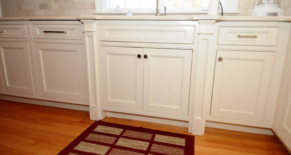 White kitchen cabinets with brushed metal handles beneath a kitchen sink, set against a light-colored countertop. A red and beige checkered rug is placed on the wooden floor in front of the cabinets.