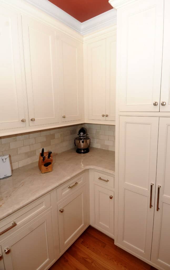 A corner kitchen with white cabinets, silver handles, light stone countertops, a knife block, a stand mixer, a white backsplash, and wooden flooring.