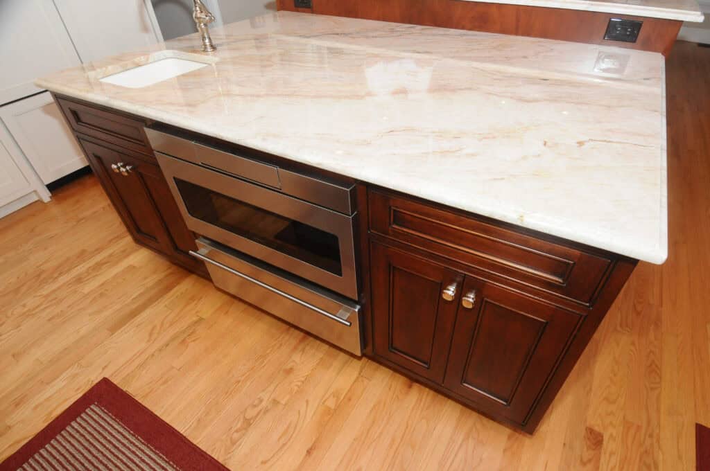 Kitchen island with a light granite countertop, stainless steel oven, dark wooden cabinets with silver handles, and a small sink in the corner, set on a wooden floor.