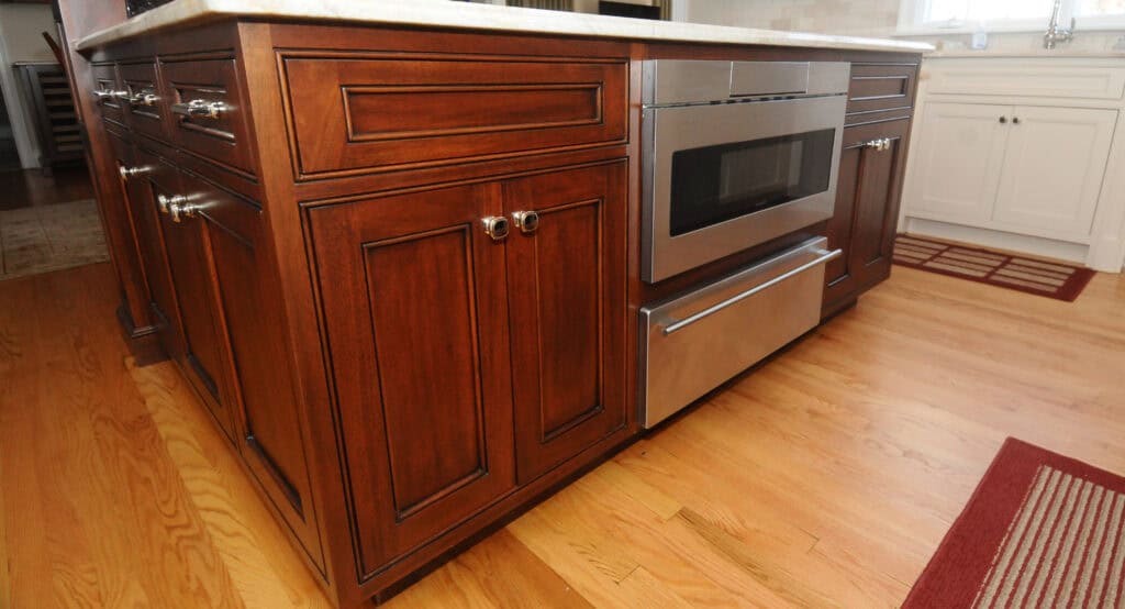 A kitchen island with dark wood cabinets and silver handles, featuring a built-in stainless steel microwave and warming drawer. The floor is light wood, and white cabinetry is visible in the background.