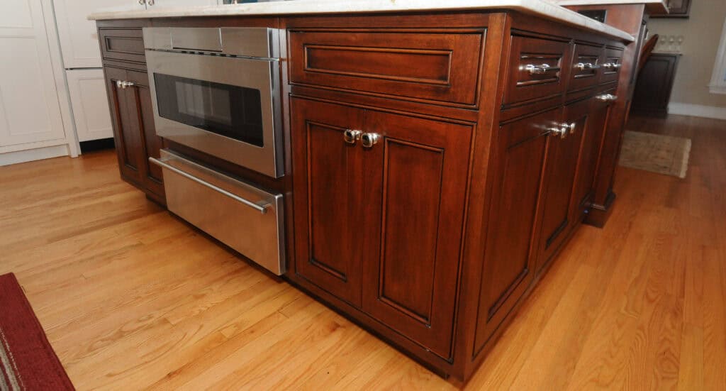 A stainless steel microwave built into a kitchen island with dark wooden cabinets and drawers, set on a light hardwood floor.