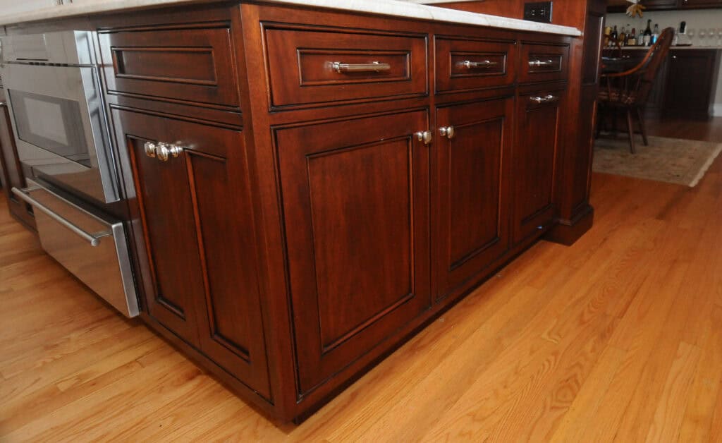 A close-up view of dark wood kitchen cabinets with silver knobs, located beneath a light countertop; stainless steel appliances and a wooden floor are also visible.