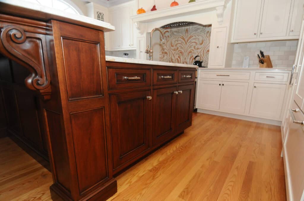 A kitchen with light wood floors, white cabinets, and a dark wood island with silver handles. Decorative corbels support the islands countertop, and there are patterned tiles behind the sink.