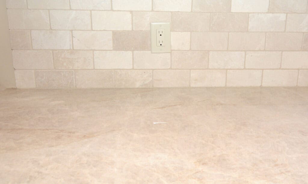 A beige stone countertop with a matching tile backsplash and a double electrical outlet centered on the wall. The surface is clean and empty.