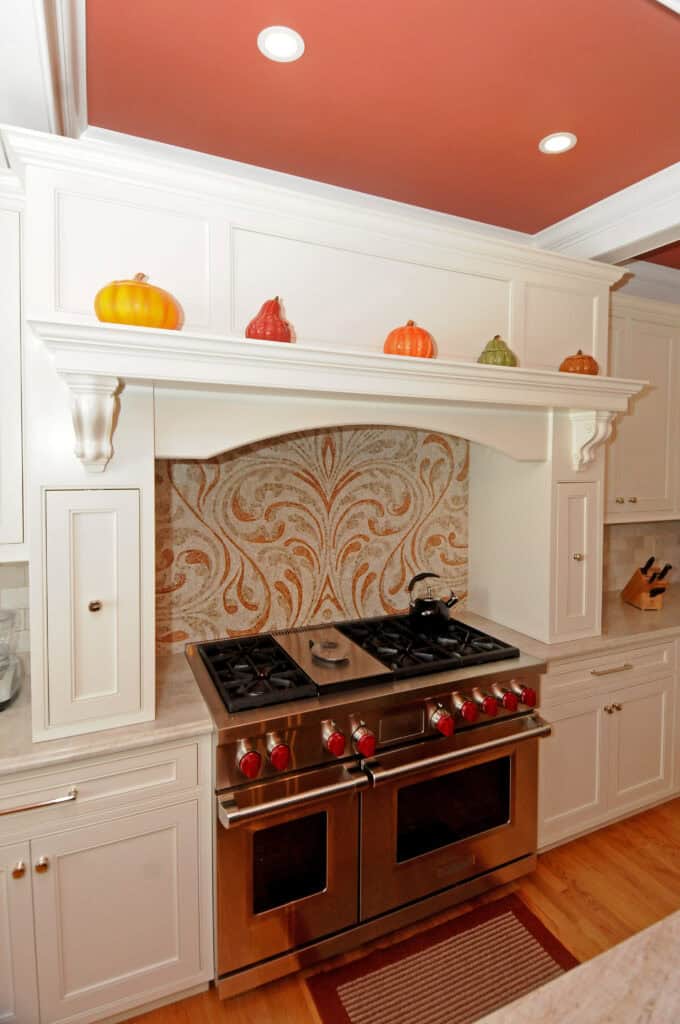 A stainless steel double oven with red knobs is set in white cabinetry, beneath a decorative range hood. The backsplash features a beige and brown paisley pattern. Ceramic pumpkins decorate the hood shelf.
