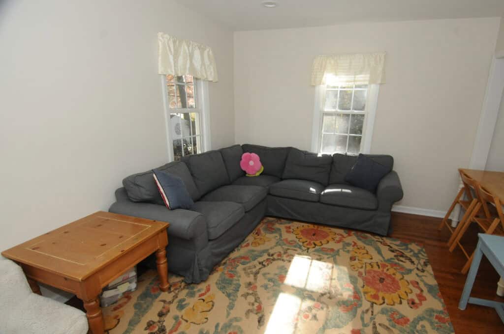 A cozy living room with a gray L-shaped sofa, colorful pillows, a floral rug, a wooden coffee table, and two windows with white curtains letting in natural light. A small dining table with chairs is in the corner.