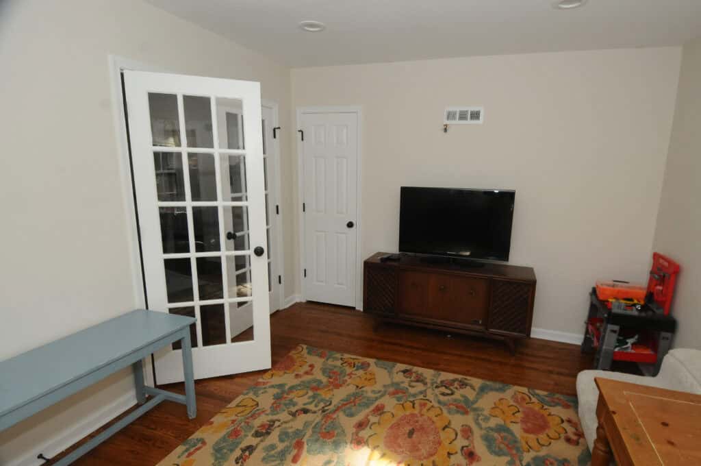 A living room with a TV on a wooden stand, a floral rug on the hardwood floor, a blue bench, a white couch, a side table, and a glass-paneled door partially open to another room.