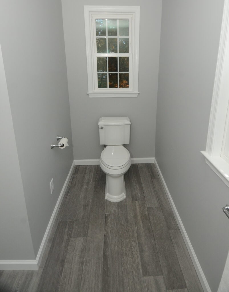 A small, modern bathroom with gray walls, wood-look tile flooring, a white toilet, a roll of toilet paper on the left wall, and a window above the toilet showing greenery outside.