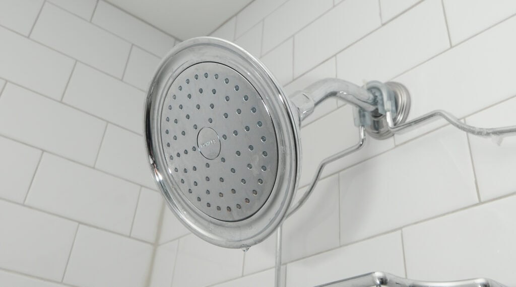 Close-up of a silver showerhead attached to a white tiled bathroom wall, with water droplets visible on its surface.