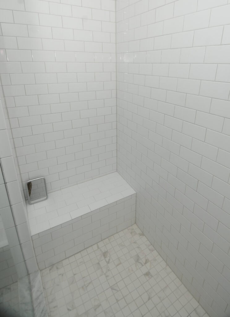 A clean, white-tiled shower with a built-in bench in the corner. The walls and bench are covered with glossy rectangular tiles, and the floor has square tiles with light gray veining.