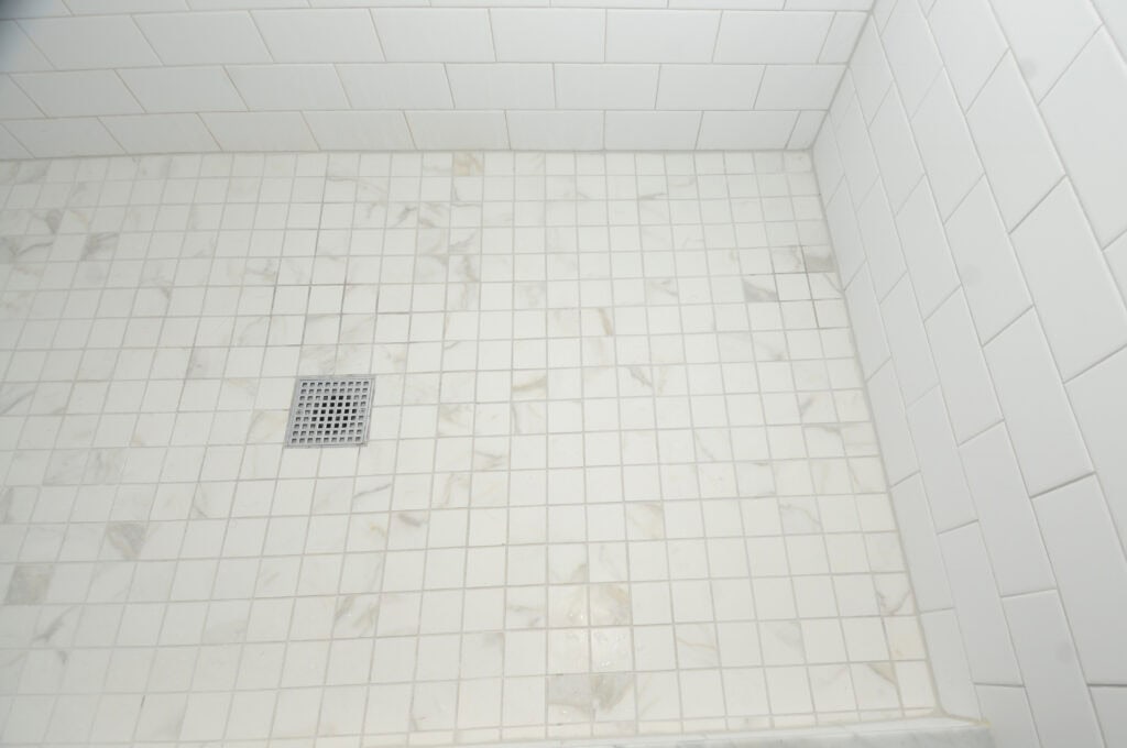 White tiled shower floor with a square metal drain cover, surrounded by white tiled walls. The tiles have a subtle marble pattern.