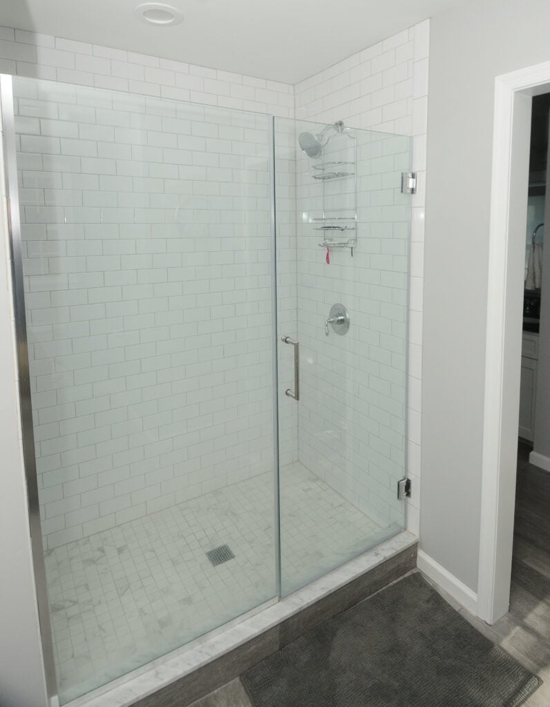 A modern bathroom shower with white subway tile walls, a glass door, chrome fixtures, and a metal shower caddy. The shower floor is tiled, and a gray bath mat is placed outside.