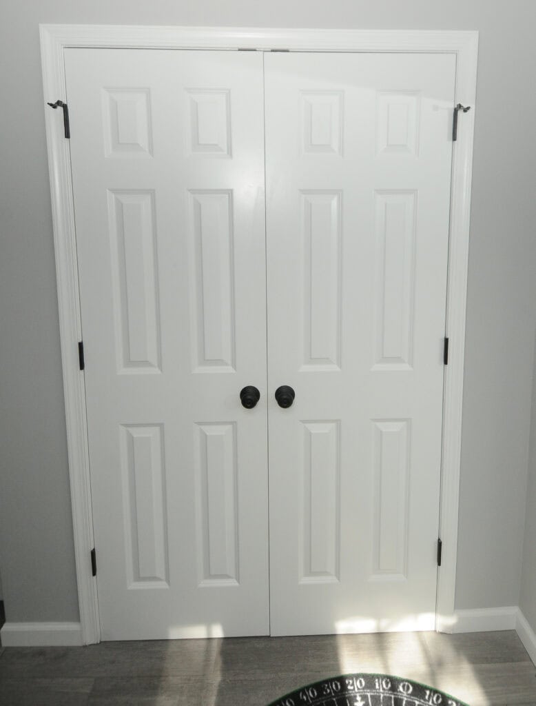 A pair of closed white double doors with black round handles, set in a light gray wall with white trim, and gray wood flooring beneath. Sunlight casts shadows near the bottom right corner.