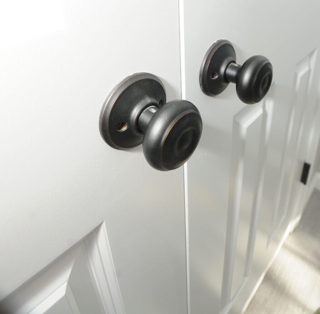 Close-up of two black round door knobs on white paneled double doors, with sunlight casting shadows across the surface.