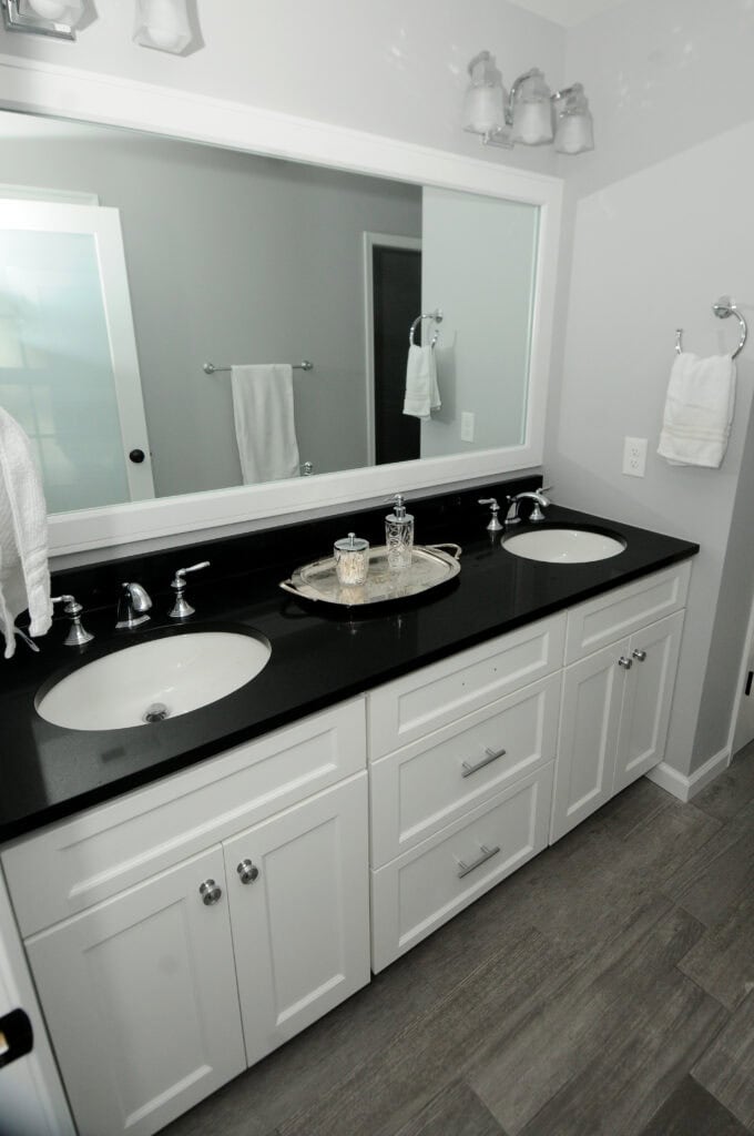 A modern bathroom with a double sink vanity, black countertop, white cabinets, large mirror, silver faucets, towel racks on the wall, and a tray with toiletries on the counter.