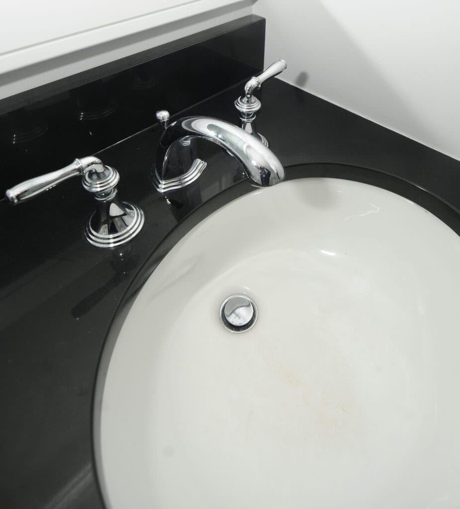 A white bathroom sink with a shiny silver faucet and handles, set in a glossy black countertop, is shown from above. The sink has some light stains near the drain.