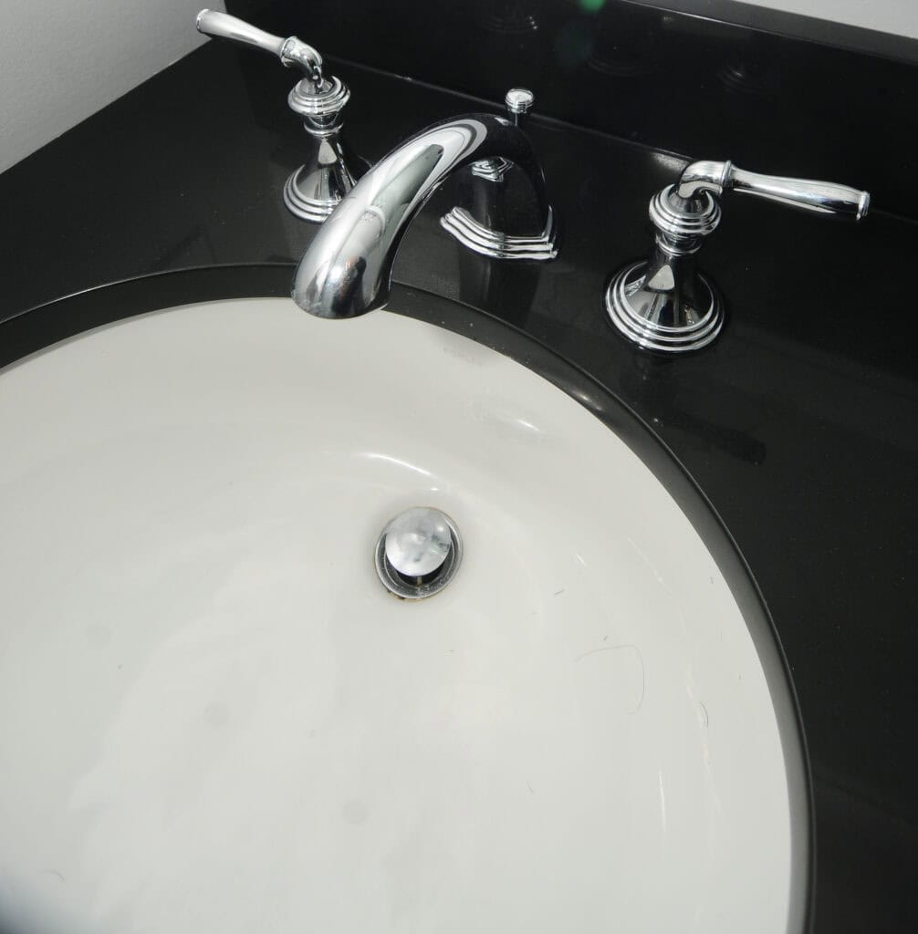 A white oval bathroom sink with a chrome faucet and two handles is set in a glossy black countertop. Some small hair strands are visible inside the sink basin.