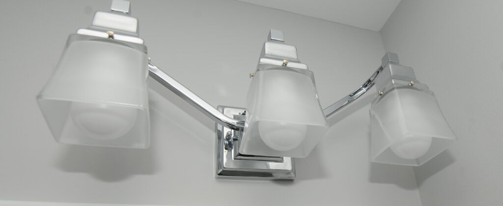 A close-up view of a modern chrome bathroom light fixture with three frosted glass square lamps mounted on a light gray wall.