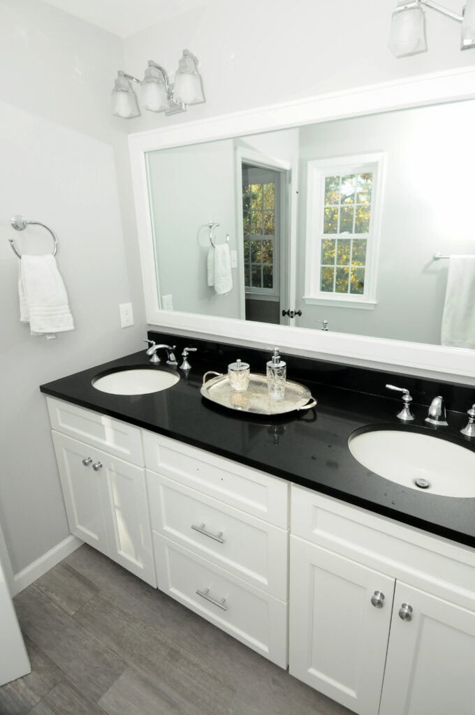 A modern bathroom with a black countertop, two white sinks, white cabinets, a large mirror, wall-mounted lights, a silver tray with glass jars, and a towel hanging on the wall near a window.