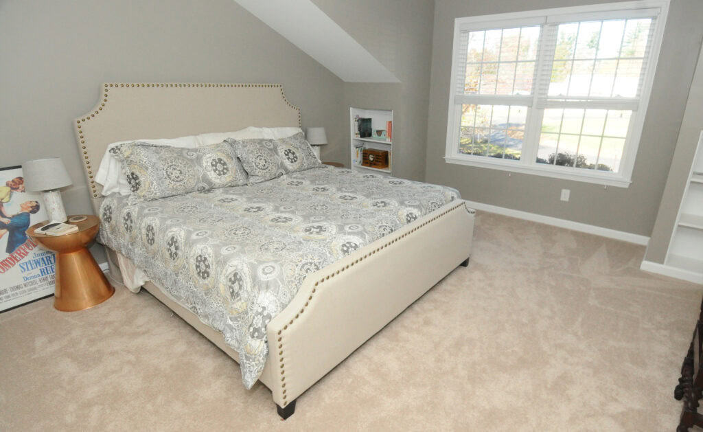 A cozy bedroom with a beige upholstered bed, patterned bedding, two nightstands with lamps, a window letting in daylight, beige carpet, and a bookshelf. A framed poster rests against the wall near the bed.