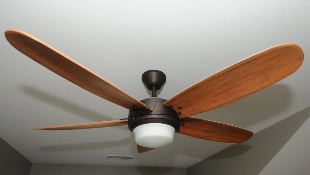 A ceiling fan with four wooden blades and a central light fixture is mounted on a white ceiling. The blades are brown, and the fan has a dark metal base.