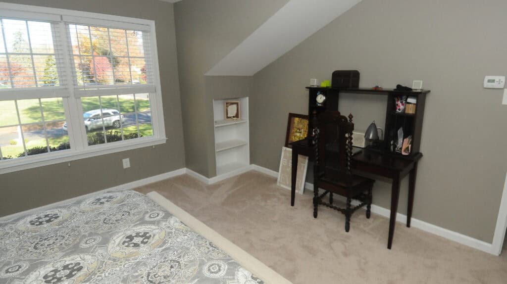 A bedroom with a bed in the foreground, a window with outdoor view, beige carpet, gray walls, a small black desk with a chair, shelves, and framed pictures resting against the wall.