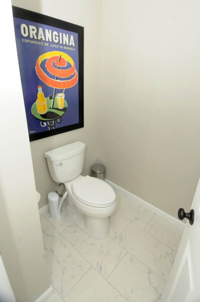 A small bathroom with a white toilet, marble-patterned floor tiles, a metal trash can, and a framed Orangina poster with colorful bottles and a glass on the wall above the toilet.