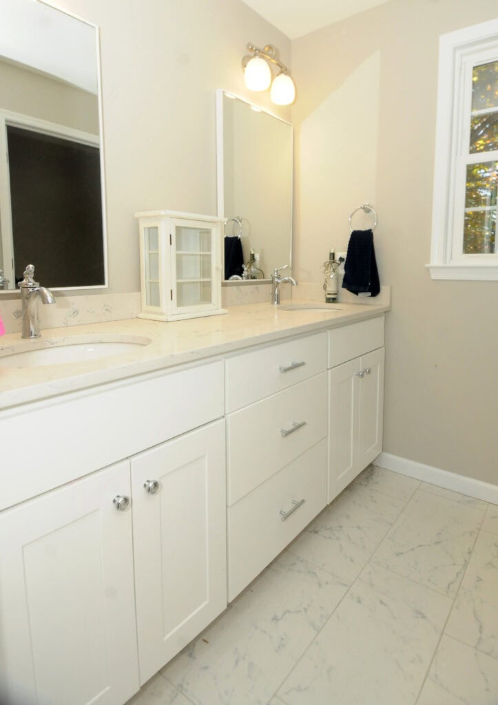 A clean, modern bathroom with white cabinets, marble countertop, dual sinks, large mirrors, and a window letting in natural light. A small glass display case and navy towel are on the counter.