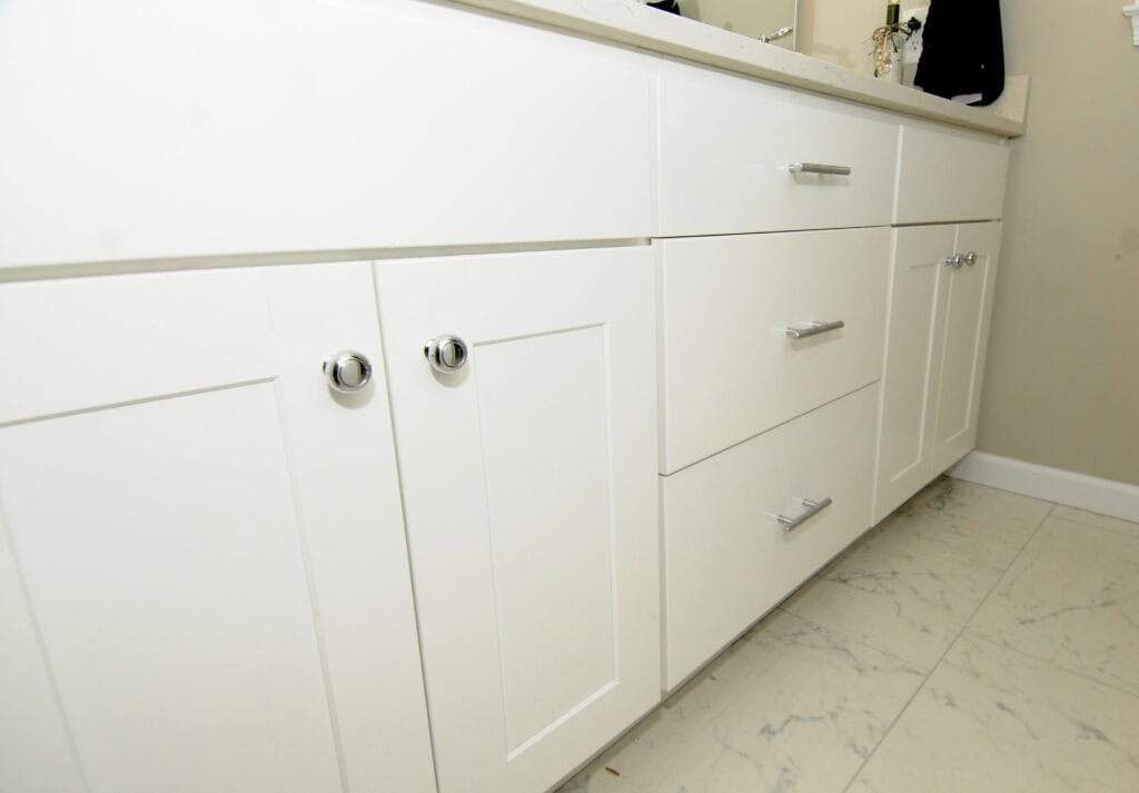 Close-up of white bathroom cabinets with silver handles and knobs, set against a light-colored marble tile floor. The countertop is partially visible at the top of the image.