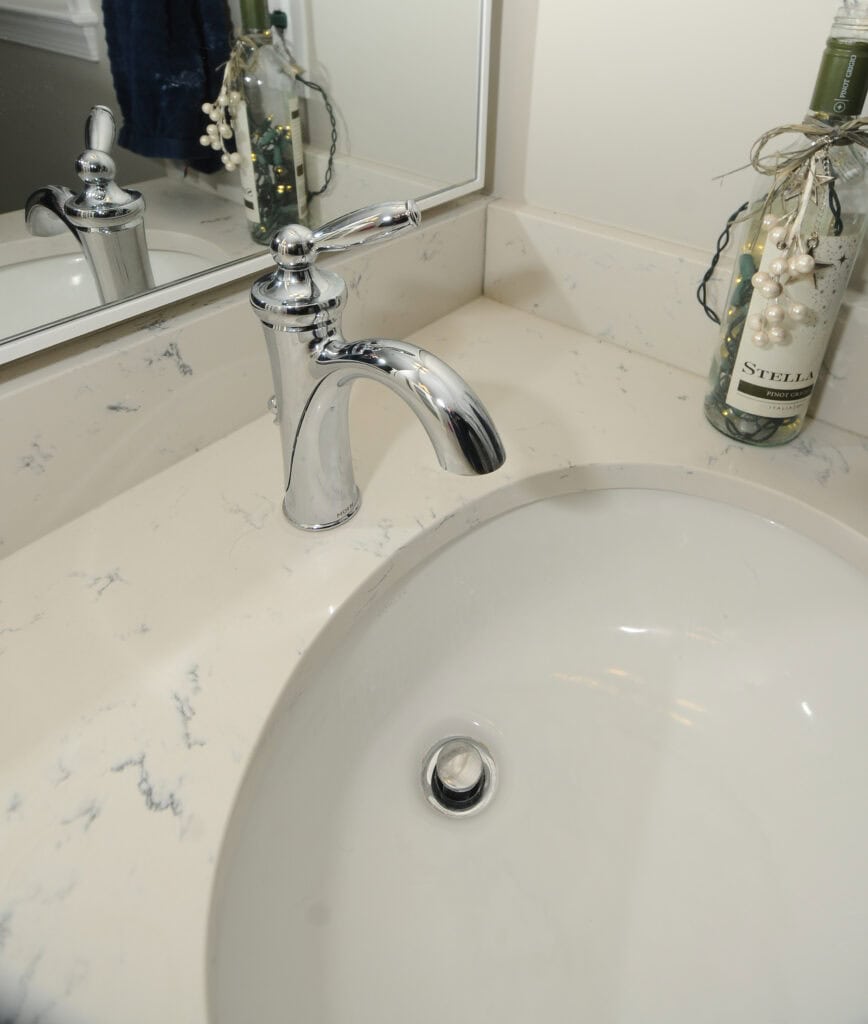A close-up view of a bathroom sink with a silver faucet, marble countertop, and a decorative glass bottle with twine and faux pearls next to a wall mirror.