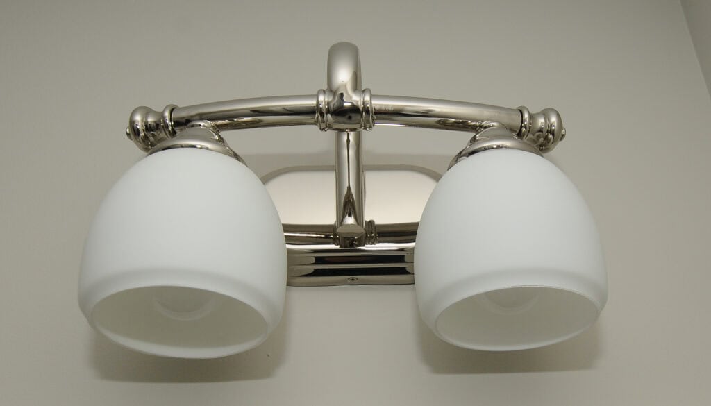 A close-up view of a modern bathroom light fixture with two frosted glass shades and a polished metal base mounted on a light-colored wall.