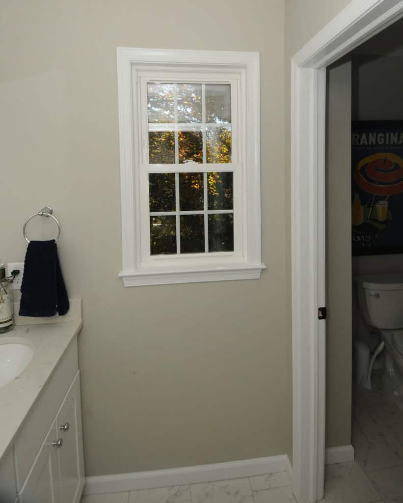 A bathroom with a white-framed window, a countertop with a towel and toiletries on the left, light beige walls, and an open door leading to a toilet in an adjacent room.