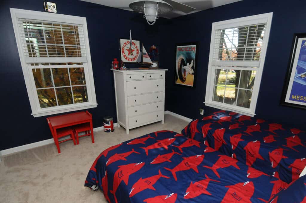 A navy blue children’s bedroom with two beds covered in red shark-patterned bedding, a white dresser, a red table with chairs, and nautical-themed decor. Three windows let in natural light.