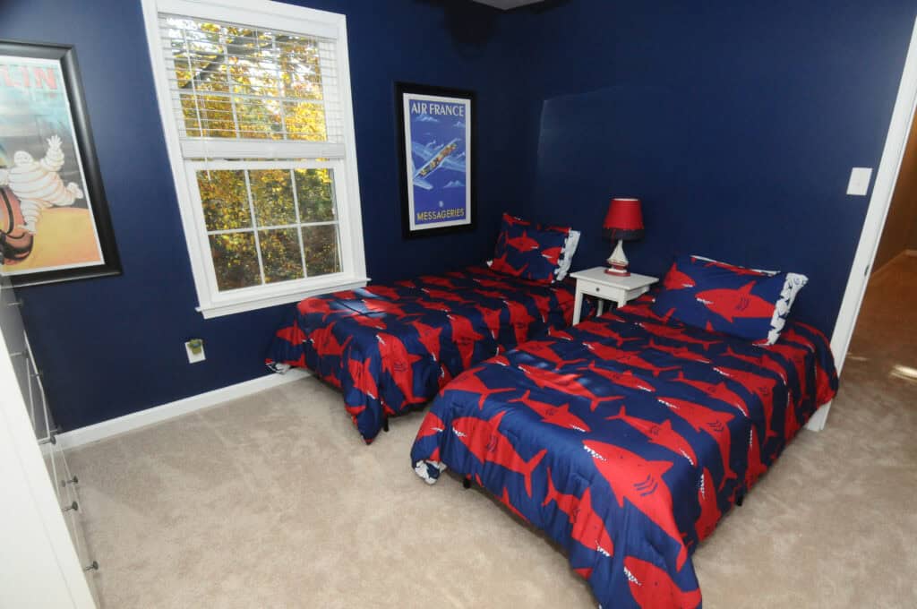 A bedroom with two twin beds covered in blue and red shark-patterned bedding, a white nightstand with a red lamp between them, dark blue walls, and a window letting in sunlight. Posters hang on the walls.