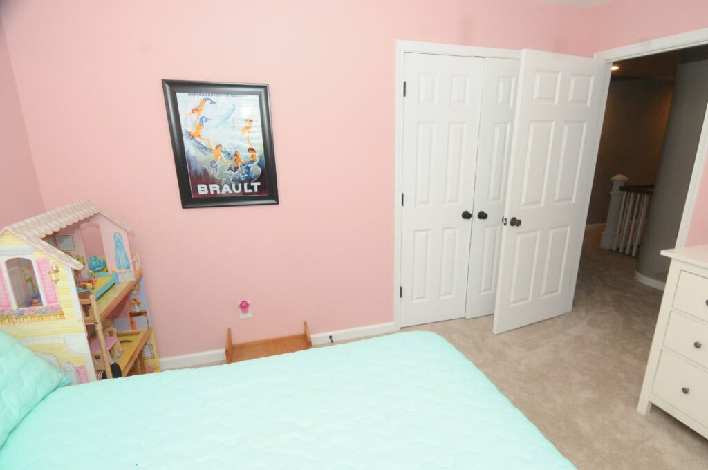 A pink-walled bedroom with a mint green bedspread, a large dollhouse, a dresser, white double doors, and a framed poster on the wall. The floor is carpeted in beige.