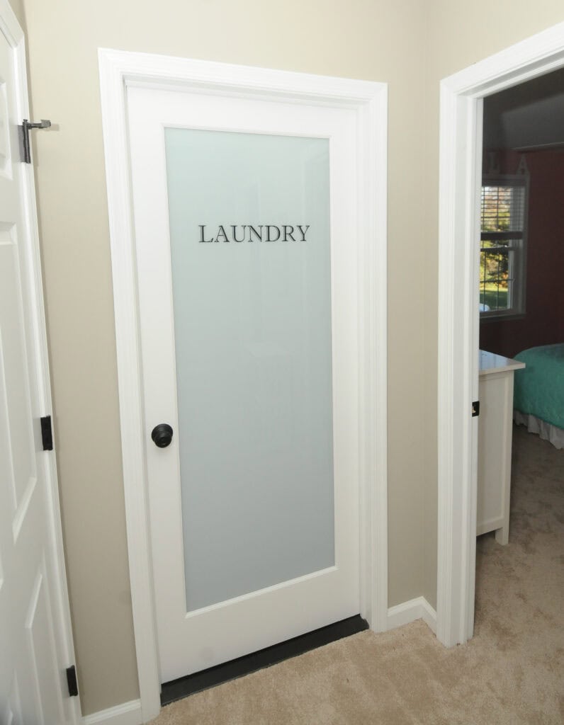 A white door with frosted glass labeled LAUNDRY in black letters, set in a hallway with beige walls and carpet. Adjacent rooms with white doors and visible bedroom furniture are nearby.