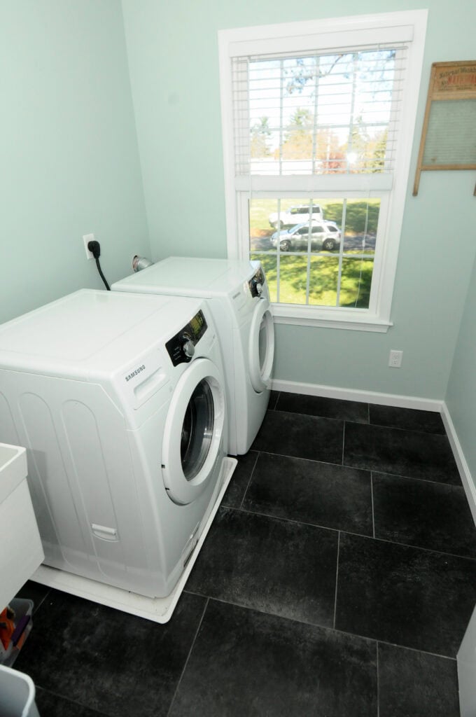 A laundry room with a washing machine and dryer side by side below a window; the room has light blue walls, black tile floor, and a washboard hanging on the wall.