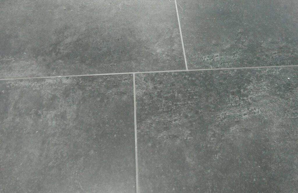 Close-up of gray stone tiles with subtle speckles and natural variations in color. The tiles are separated by thin, light-colored grout lines forming a grid pattern.