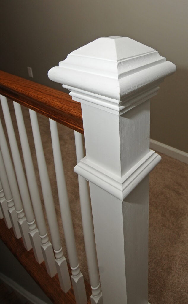 A close-up of a white wooden newel post at the end of a staircase railing with white balusters and a wooden handrail, set against a beige carpeted floor and neutral wall.