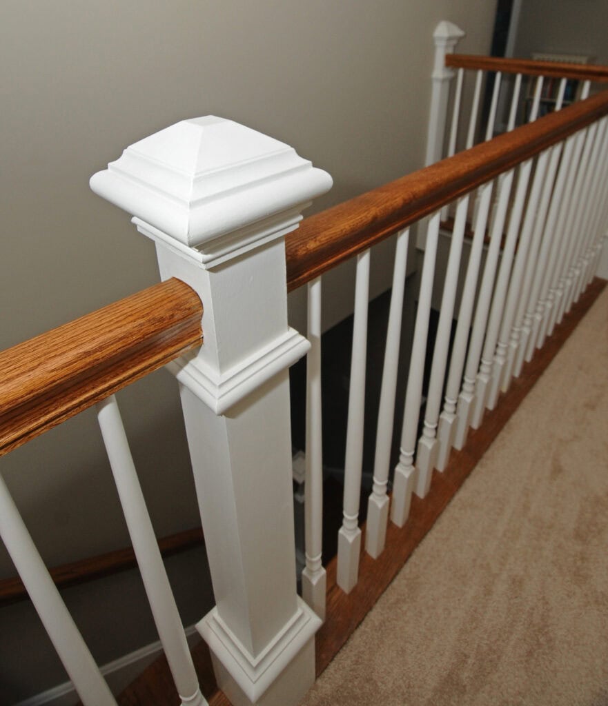 A close-up view of a staircase railing with a wooden handrail, white spindles, and a decorative square newel post on a carpeted floor. The wall and stair area are painted in neutral tones.